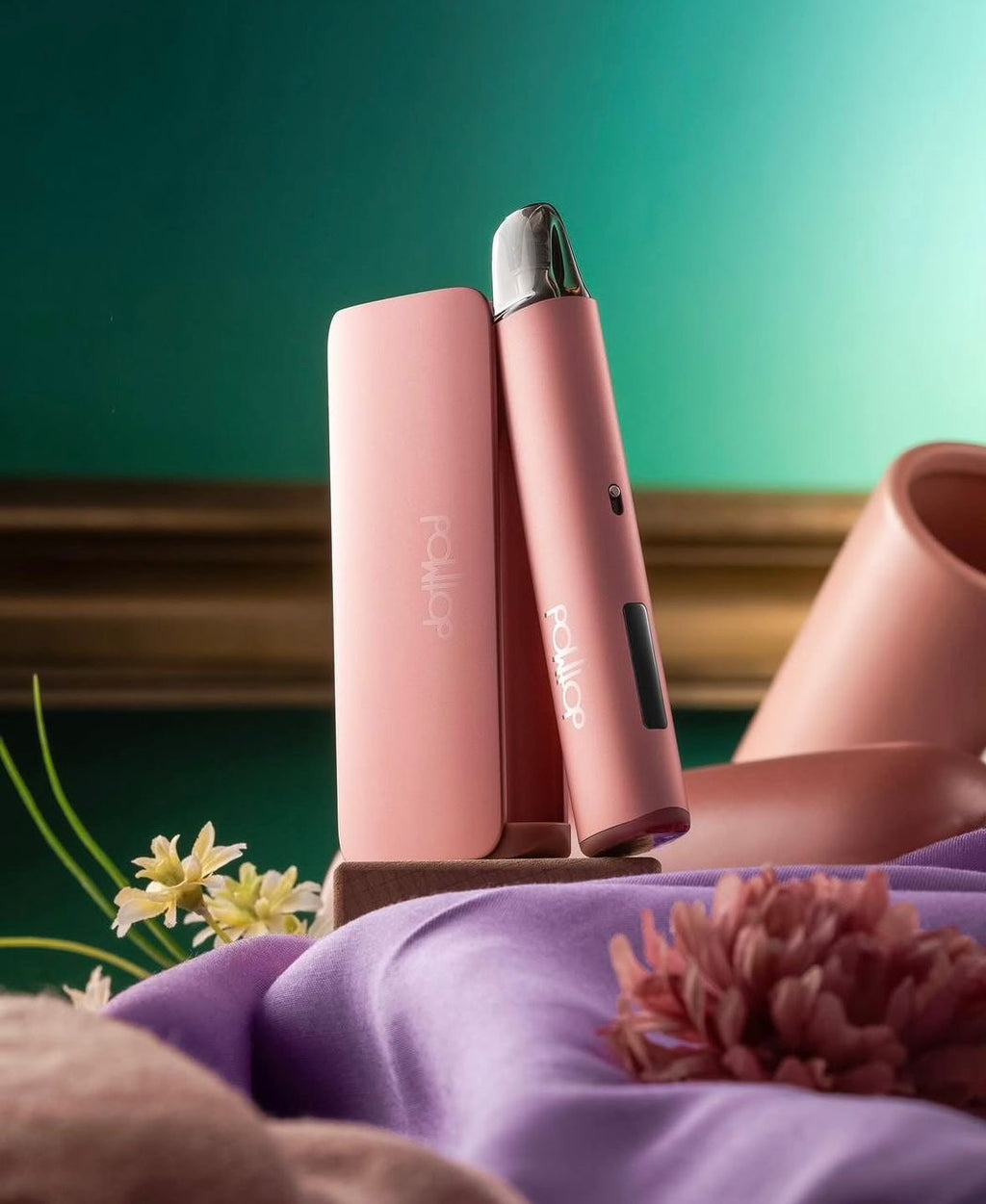 Pink DottMod vape device with charging case, styled with flowers on purple fabric against teal background