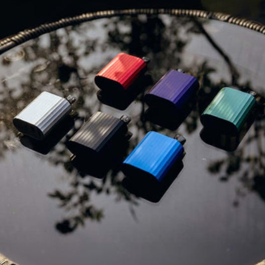 Six colorful vape pods arranged on reflective surface, showcasing red, blue, green, silver, black, and purple options